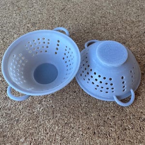 May include: Two light blue plastic colanders with handles. The colanders are round with many small holes for draining. One colander is upright, and the other is turned upside down. The colanders are kitchen tools.