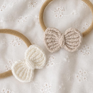 May include: Two hair headbands featuring crocheted bows. One headband has a cream-colored bow, and the other has a beige-colored bow. Both headbands are made of a tan-colored elastic material, providing a simple and functional design for hair.