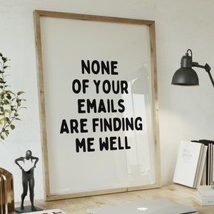 以下が含まれることがあります： 白い壁に「None of your emails are finding me well」というテキストが書かれた額縁付きのプリントが飾られています。その下には、ラップトップ、本、ランプ、ムーンランプが置かれた木製デスクがあります。