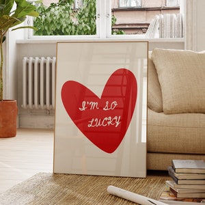 May include: A framed print with a red heart and the text "I'm so lucky" in white. The print is hanging on a white wall in a living room with a couch, a chair, and a plant.