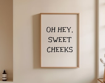 May include: A framed print with the text "Oh hey, sweet cheeks" hanging on a white wall in a bathroom. The bathroom has a white bathtub, a wooden floor, and a window with a vase of dried flowers.