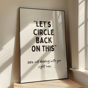 May include: A white framed print with black text that reads "Let's Circle Back On This" and "100% not dealing with you right now." The print is hanging on a white wall next to a window.