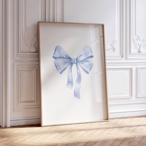 May include: A framed print of a watercolor painting of a large blue bow. The bow is tied in a knot and the ends of the ribbon are flowing down. The print is hanging on a white wall in a room with white trim and wood floors.