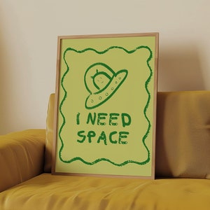 Könnte beinhalten: Ein gerahmter Druck mit einer grünen Alien-Zeichnung und dem Text "I NEED SPACE" auf gelbem Hintergrund.