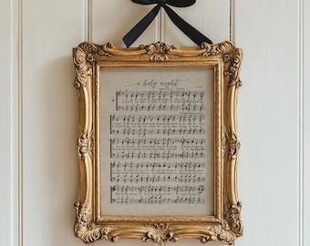 May include: A framed print of sheet music with the words "o holy night" in elegant script. The print is in a gold ornate frame, suspended by a black ribbon against a white paneled wall. The frame has a vintage, antique aesthetic.
