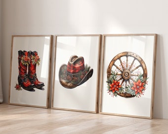 May include: Three framed watercolor prints featuring a Western theme. The prints depict red cowboy boots with floral accents, a plaid cowboy hat, and a wooden wagon wheel with floral and berry decorations. The frames are light-colored wood.