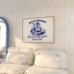 May include: A framed print with the text "READ BOOKS" and "Because who needs social life?" in blue. The image features a vintage illustration of a woman reading. The print is in a wooden frame and hangs on a white paneled wall.