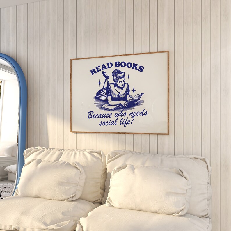 May include: A framed print with the text "READ BOOKS" and "Because who needs social life?" in blue. The image features a vintage illustration of a woman reading. The print is in a wooden frame and hangs on a white paneled wall.