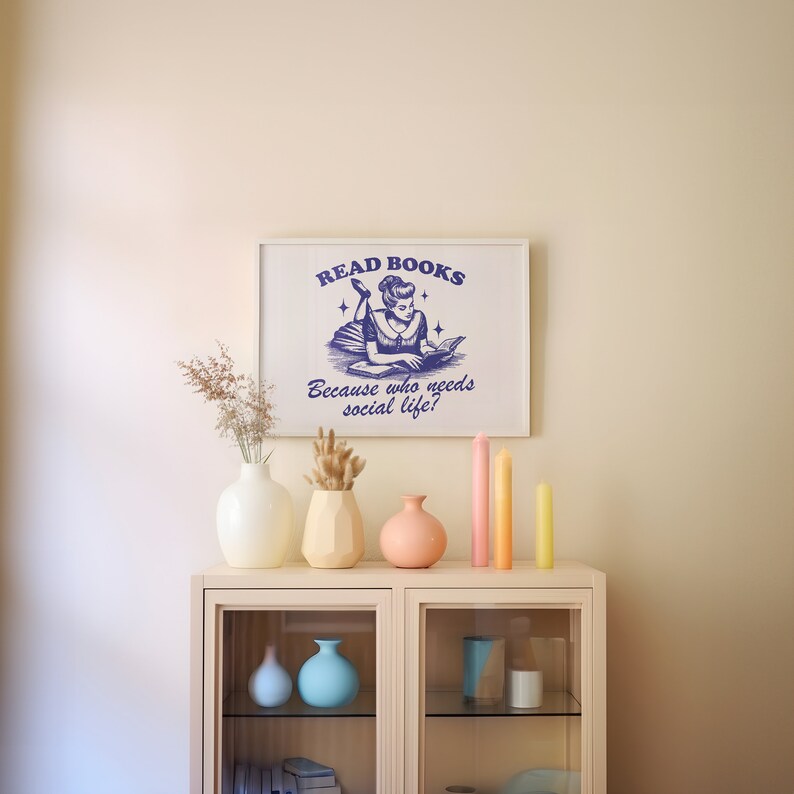 May include: A white framed print with the text "READ BOOKS Because who needs social life?" is displayed above a cabinet. The cabinet holds vases, candles, and decorative items. The print features a blue illustration of a woman reading.