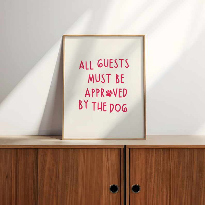 Puede incluir: Una impresi&oacute;n enmarcada con el texto "ALL GUESTS MUST BE APPROVE BY THE DOG" en letras rosas con una huella de pata en la palabra "APPROVED". El marco es de madera clara y el fondo es blanco.