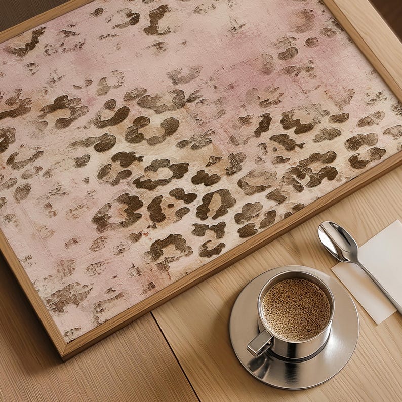 May include: A framed print featuring an abstract leopard print design in shades of pink and brown. The print is displayed on a wooden table alongside a cup of coffee and a spoon.