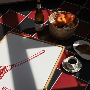 May include: A framed print with the text "NOTHING IS ASTABLE" and a red illustration of a broom. The print is on a black and red checkered floor with a bottle of wine, a basket of apples, a cup of coffee, and a croissant.
