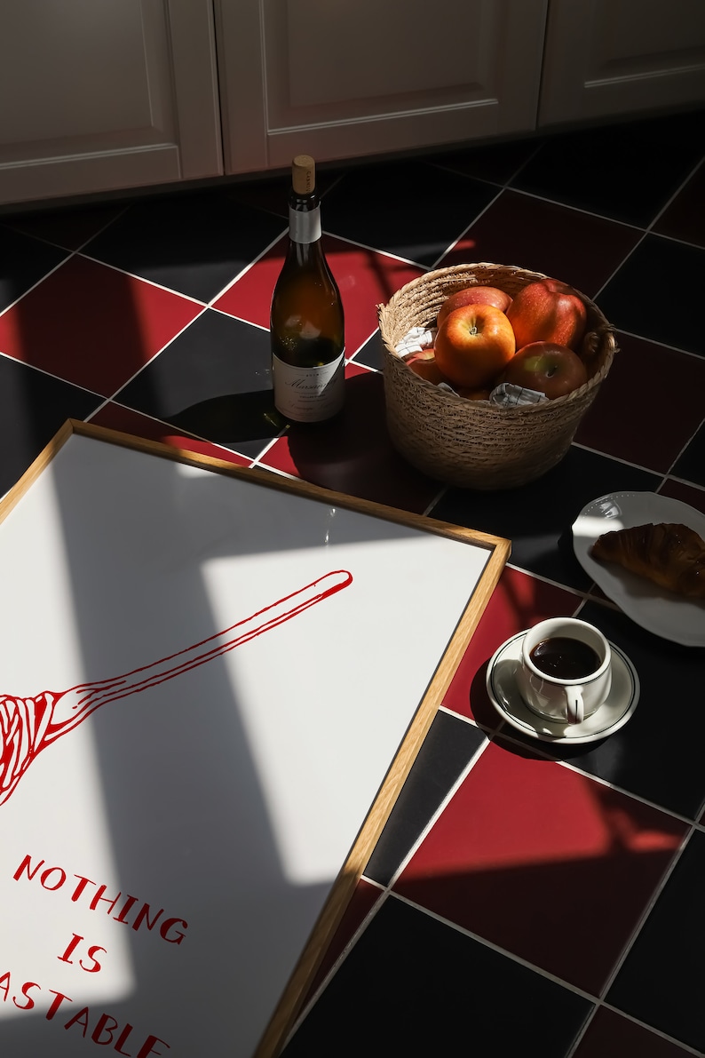 May include: A framed print with the text "NOTHING IS ASTABLE" and a red illustration of a broom. The print is on a black and red checkered floor with a bottle of wine, a basket of apples, a cup of coffee, and a croissant.