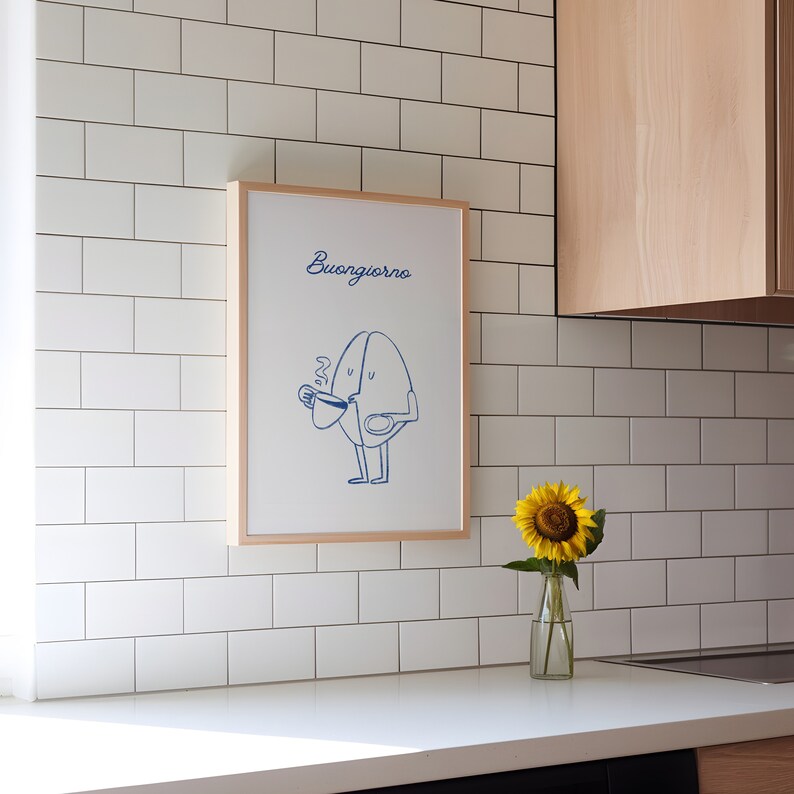 Peut inclure: Une impression encadr&eacute;e avec un fond blanc et une illustration bleue d'un grain de caf&eacute; avec un visage et des bras tenant une tasse de caf&eacute;. Le texte "Buongiorno" est &eacute;crit en bleu en haut de l'impression.