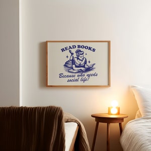 May include: A framed print with the text "READ BOOKS Because who needs social life?" in navy blue. The illustration depicts a woman reading a book. The frame is made of light brown wood. The print is hanging on a white wall.