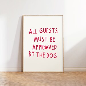 Puede incluir: Un cartel enmarcado blanco con el texto "ALL GUESTS MUST BE APPROVED BY THE DOG" en letras rojas con una huella de pata roja en la palabra "APPROVED".