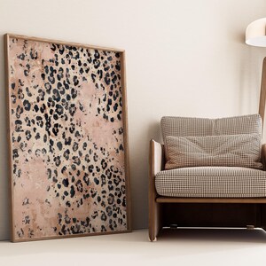 May include: A large framed print with a pink and black leopard print pattern. The print is hanging on a light beige wall next to a brown armchair with a checkered fabric.