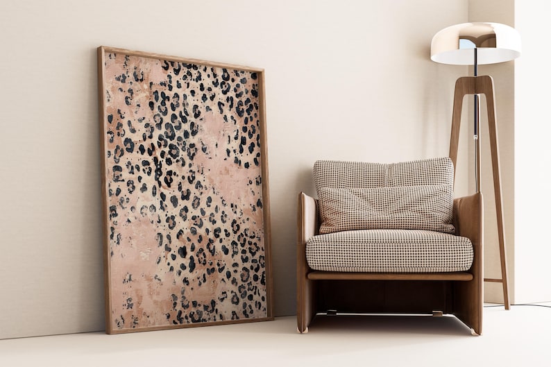 May include: A large framed print with a pink and black leopard print pattern. The print is hanging on a light beige wall next to a brown armchair with a checkered fabric.