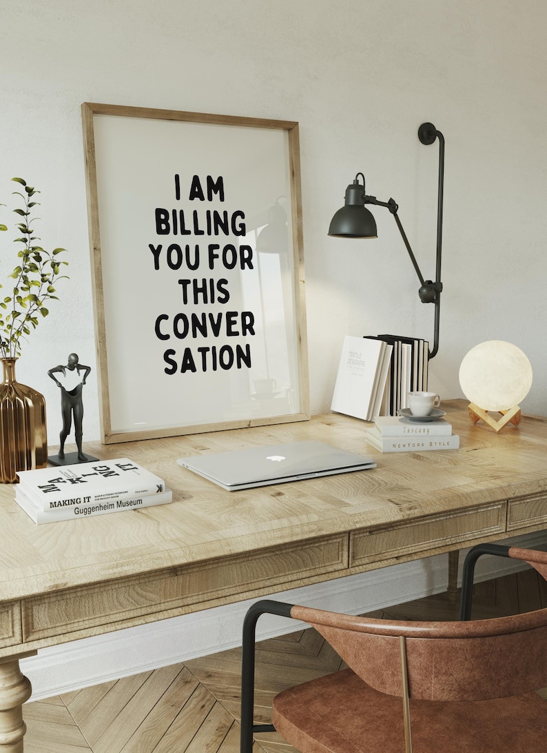 May include: A framed print with the text "I AM BILLING YOU FOR THIS CONVERSATION" in black lettering on a white background. The print is on a wooden desk with a laptop, books, a lamp, and a moon-shaped lamp.