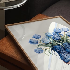 K&ouml;nnte beinhalten: Ein gerahmtes Aquarellbild von blauen Tulpen in einem Paar blauer Stiefel. Das Bild h&auml;ngt auf einem Holztisch.