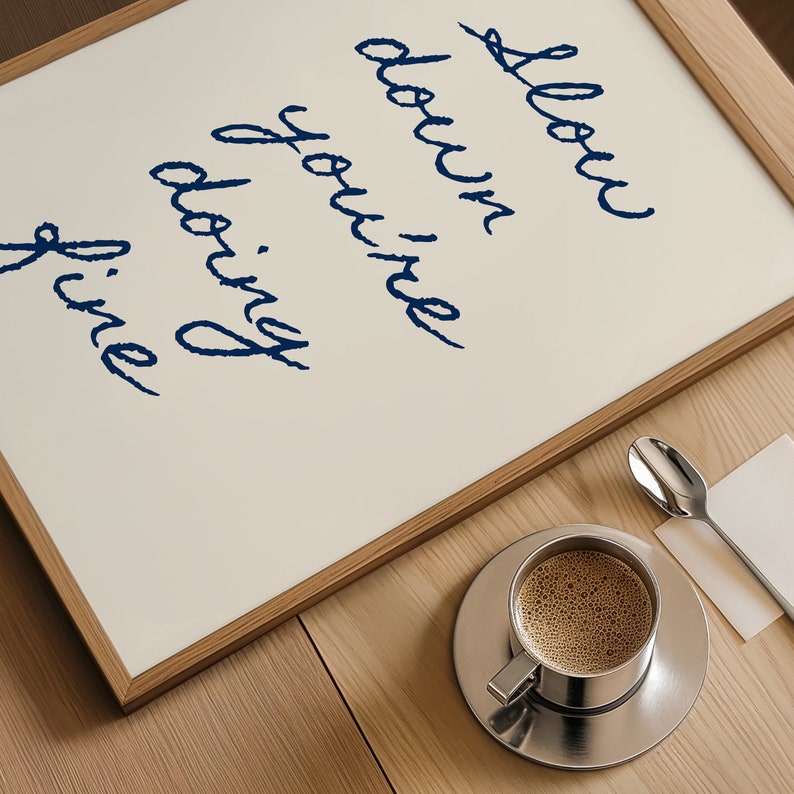 May include: A framed print with the text "Slow down you're doing fine" in blue cursive writing. A cup of coffee and a spoon are on a wooden table in the foreground.