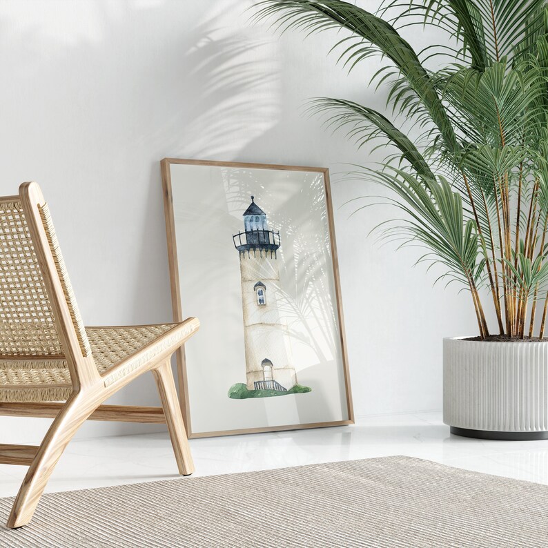 May include: A watercolor painting of a white lighthouse with a blue roof and a green grassy base. The painting is framed in a wooden frame and is hanging on a white wall. A wicker chair and a potted plant are in the foreground.