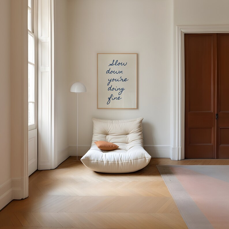 May include: A white armchair with a brown throw pillow in a room with a framed print that reads "Slow down you're doing fine".