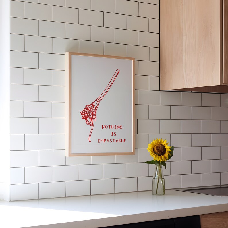 May include: A white framed print with a red illustration of a bundle of spaghetti and the text "Nothing is Impassable". A sunflower in a glass vase sits on a white countertop in front of the print.