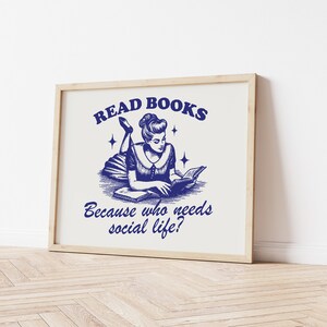 May include: A framed print featuring a blue ink illustration of a woman reading a book. The text reads "READ BOOKS Because who needs social life?" The artwork is set against a cream background and is in a light wooden frame.