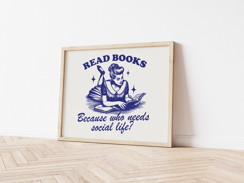 May include: A framed print featuring a blue ink illustration of a woman reading a book. The text reads "READ BOOKS Because who needs social life?" The artwork is set against a cream background and is in a light wooden frame.