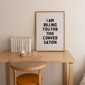 May include: A framed print with the text "I AM BILLING YOU FOR THIS CONVERSATION" in black lettering on a white background. The print is hanging on a white wall above a wooden desk with a chair and books.