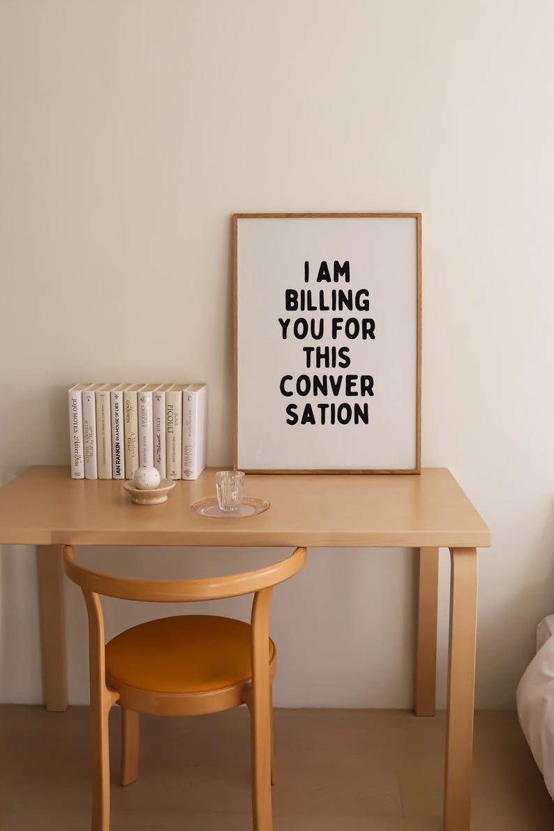 May include: A framed print with the text "I AM BILLING YOU FOR THIS CONVERSATION" in black lettering on a white background. The print is hanging on a white wall above a wooden desk with a chair and books.