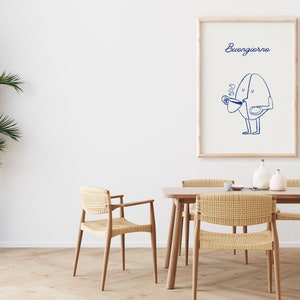 Peut inclure: Une impression encadr&eacute;e avec un dessin au trait bleu d'un personnage en forme de grain de caf&eacute; tenant une tasse de caf&eacute;. Le texte "Buongiorno" est &eacute;crit au-dessus du personnage. L'impression est accroch&eacute;e &agrave; un mur blanc au-dessus d'une table en bois avec des chaises en osier.
