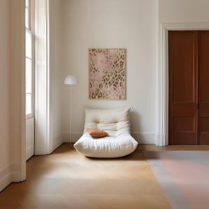 May include: A white, tufted, oversized chair with a brown cushion sits in a room with white walls and wooden floors. A framed print with a pink and brown abstract pattern hangs on the wall behind the chair.