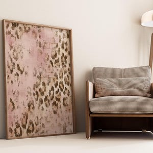 May include: A framed print of a pink and brown abstract leopard print design. The print is hanging on a white wall next to a brown armchair with a checkered pattern.