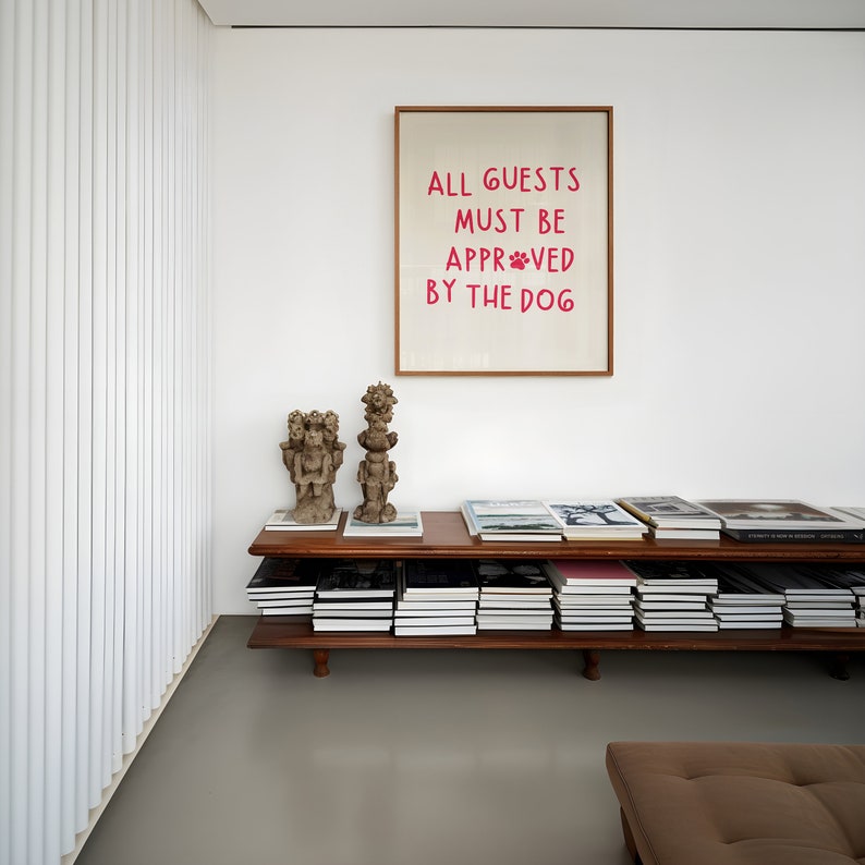 Puede incluir: Una estanter&iacute;a de madera con dos estantes, llena de libros. Dos peque&ntilde;as esculturas decorativas marrones est&aacute;n en el estante superior. Una impresi&oacute;n enmarcada con el texto "ALL GUESTS MUST BE APPROVED BY THE DOG" cuelga en la pared sobre la estanter&iacute;a.