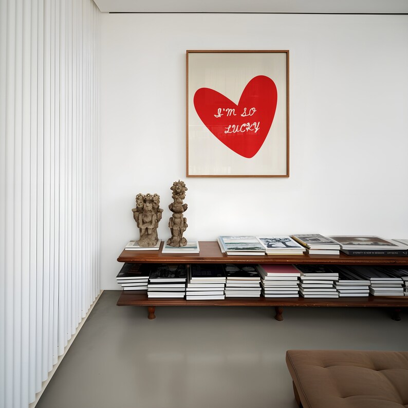 May include: A wooden shelf unit with two shelves, filled with books. The shelf is against a white wall with a framed print of a red heart with the text "I'm so lucky" above it. Two small decorative sculptures are on the top shelf.
