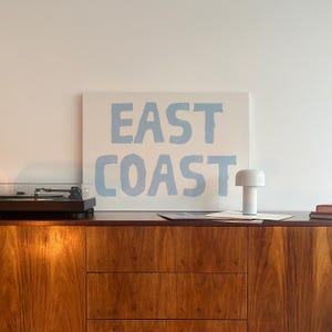 May include: A white canvas with the words "EAST COAST" printed in light blue paint. The canvas is hanging on a white wall above a wooden cabinet with a record player and a white lamp.