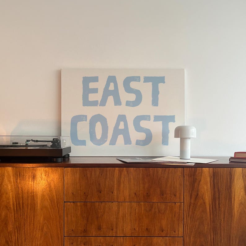 May include: A white canvas with the words "EAST COAST" printed in light blue paint. The canvas is hanging on a white wall above a wooden cabinet with a record player and a white lamp.