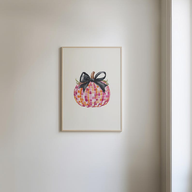 May include: A framed watercolor painting of a pink and orange checkered pumpkin with a black bow. The pumpkin is the focal point of the image and is set against a white background.