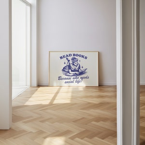 May include: A framed print with a vintage blue illustration. It depicts a woman reading a book, with the text "READ BOOKS" and "Because who needs social life?" The frame is light brown. The print is displayed in a room with wooden flooring.