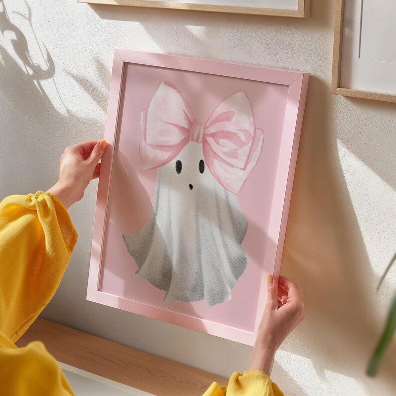 Pu&ograve; includere: Una stampa incorniciata rosa di un fantasma bianco che indossa un fiocco rosa. Il fantasma ha due occhi neri e una piccola bocca.