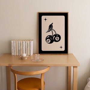 Op de afbeelding: Een zwart-witte illustratie van twee kersen met het nummer 8 op elk. De kersen bevinden zich op een lichtroze achtergrond met een zwarte lijst. De illustratie is ingelijst en hangt aan een muur boven een houten tafel.