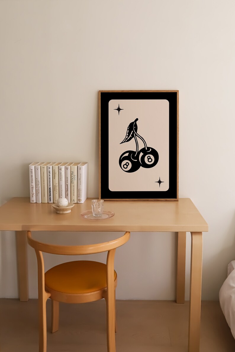 Op de afbeelding: Een zwart-witte illustratie van twee kersen met het nummer 8 op elk. De kersen bevinden zich op een lichtroze achtergrond met een zwarte lijst. De illustratie is ingelijst en hangt aan een muur boven een houten tafel.