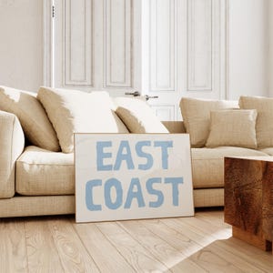 May include: A framed print with the text "EAST COAST" in light blue lettering on a white background. The print is leaning against a beige couch with pillows.
