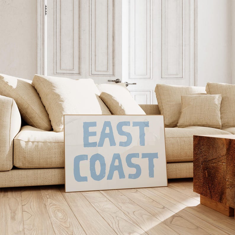 May include: A framed print with the text "EAST COAST" in light blue lettering on a white background. The print is leaning against a beige couch with pillows.
