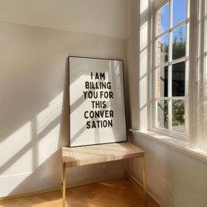May include: A black framed poster with white text that reads "I am billing you for this conversation" hanging on a white wall next to a window. A light brown wicker bench with gold legs sits in front of the poster.