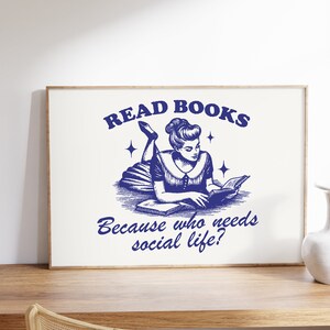 May include: A framed print with a vintage navy blue illustration. It depicts a woman reading a book, with the text "READ BOOKS" above and "Because who needs social life?" below. The print is displayed on a wooden surface.