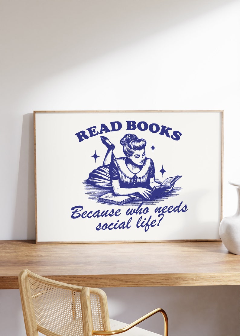 May include: A framed print with a vintage navy blue illustration. It depicts a woman reading a book, with the text "READ BOOKS" above and "Because who needs social life?" below. The print is displayed on a wooden surface.