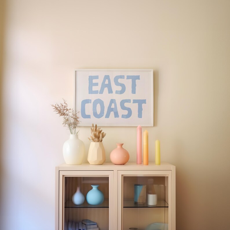 May include: A white cabinet with glass doors displays a collection of pastel-colored vases and candles. The cabinet is decorated with a framed print that reads "EAST COAST" in blue lettering.